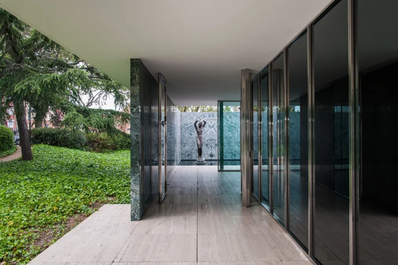 Galería exterior del Pabellón Mies van der Rohe, con vidrio, piedra y una escultura al fondo